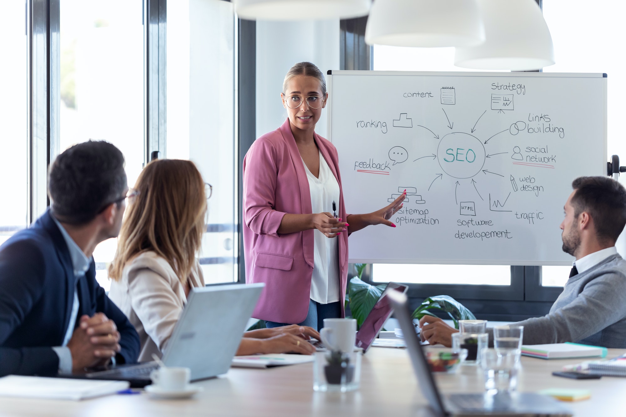 young businesswoman pointing at white blackboard and explain a project to her colleagues jpg